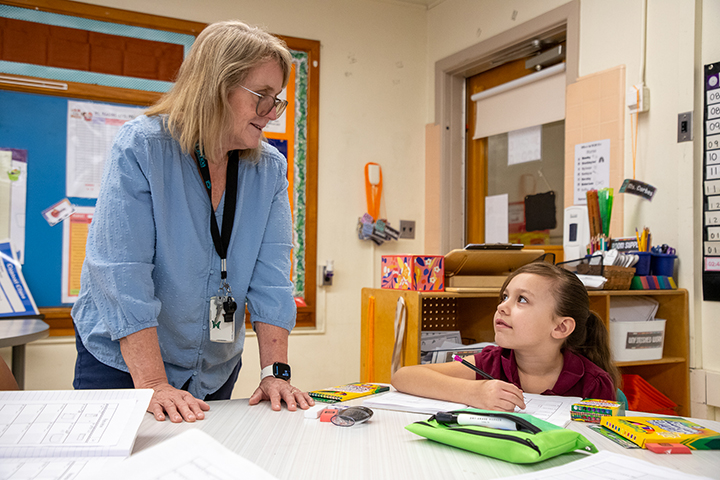 A woman in a blue shirt talks to a girl sitting at her desk