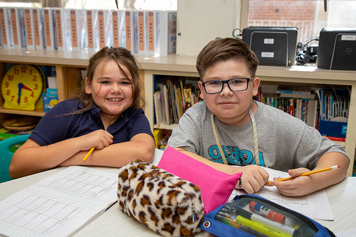 A girl and boy smile while sitting at their table in the classroom