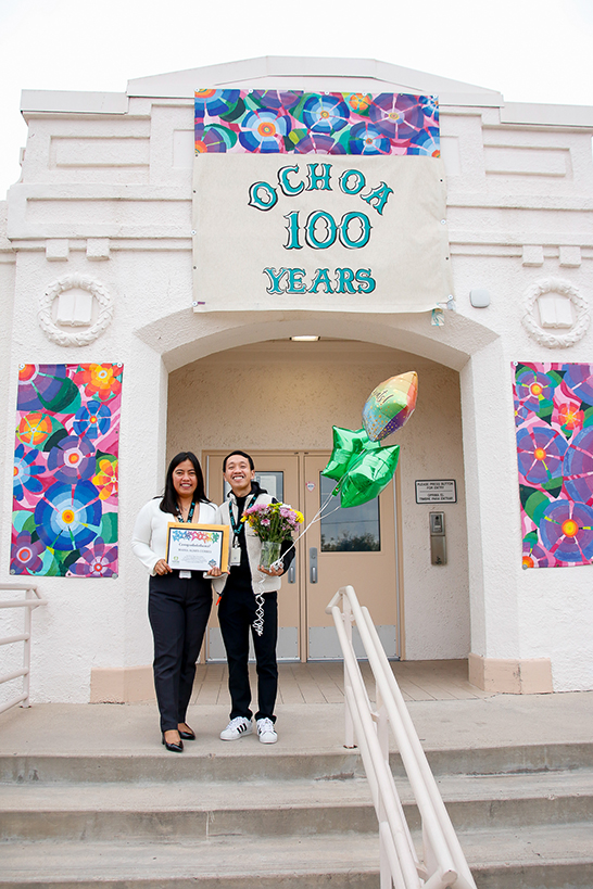 Two women stand at the top of a staircase under the Ochoa 100 Years Banner