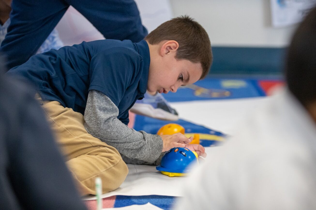 A boy in a blue shirt sits on the floor working on his STEM robot